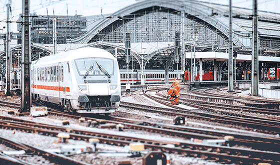 Un treno ICE bianco parte da una stazione principale. Sullo sfondo, operai che lavorano sui binari